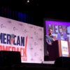 Charlie Kirk responde a preguntas de los asistentes desde el podio del escenario del “American Comeback Tour”, mientras una pantalla ampliada dividida muestra su imagen y la de un asistente del evento. Al fondo, el logo del evento.
