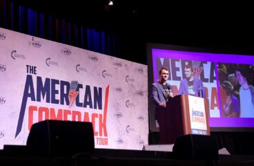 Charlie Kirk responde a preguntas de los asistentes desde el podio del escenario del “American Comeback Tour”, mientras una pantalla ampliada dividida muestra su imagen y la de un asistente del evento. Al fondo, el logo del evento.
