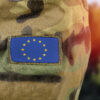 Close-up of a camouflage military uniform with a European Union flag patch sewn on the sleeve, with a blurred background in warm tones.