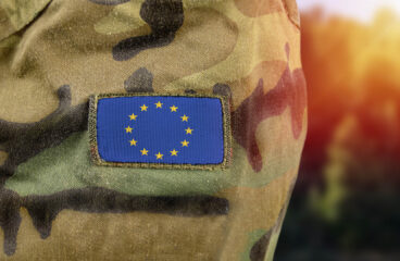 Close-up of a camouflage military uniform with a European Union flag patch sewn on the sleeve, with a blurred background in warm tones.