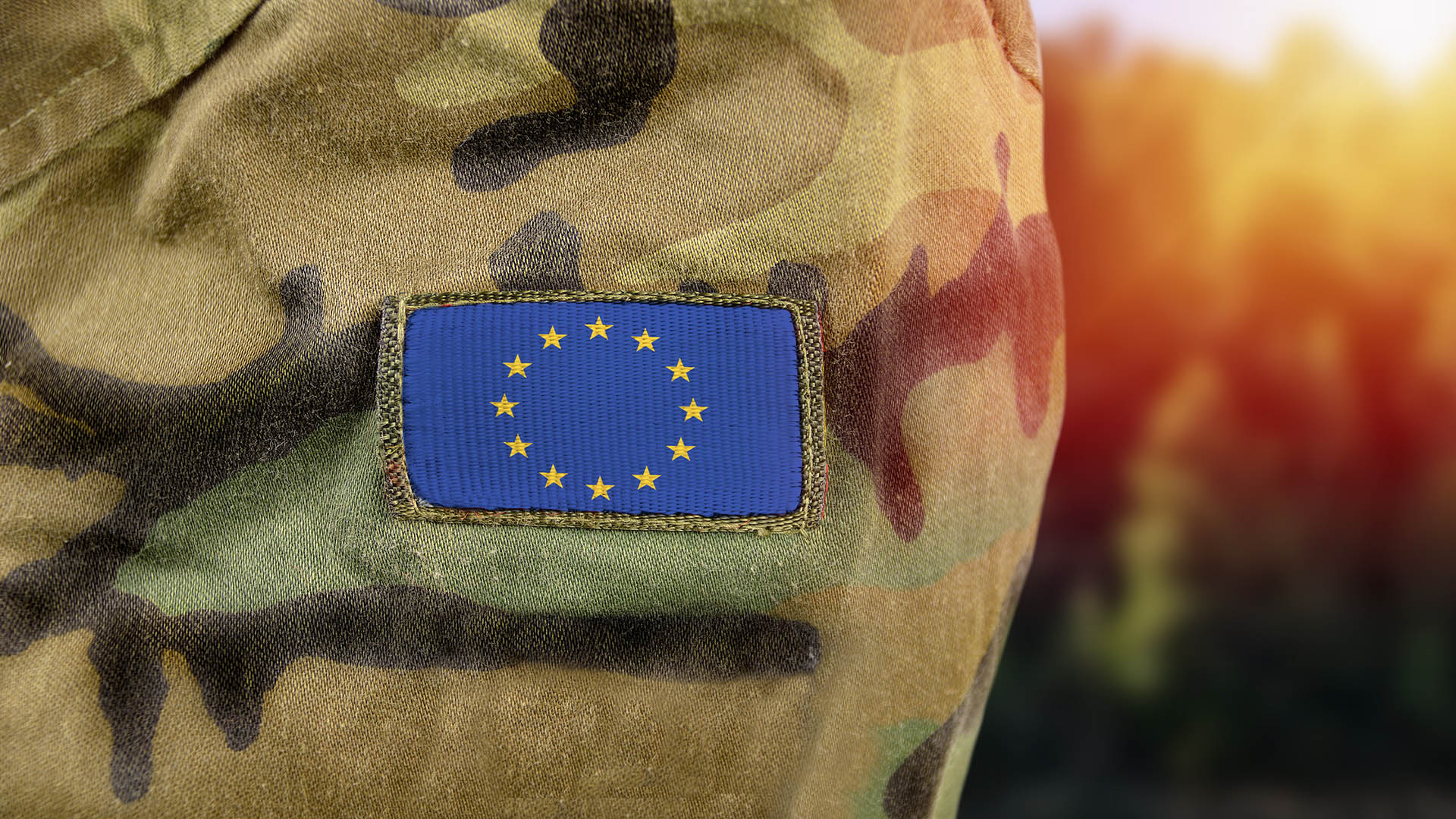 Close-up of a camouflage military uniform with a European Union flag patch sewn on the sleeve, with a blurred background in warm tones.