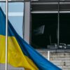 A Ukrainian flag flies from a flagpole in front of a building damaged by bombing during the Russian war in Kyiv. In the background, broken glass, walls blackened by fire and the remains of office furniture.