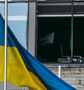 Una bandera de Ucrania ondea en un mástil frente a un edificio dañado por los bombardeos por la guerra de Rusia en Kyiv. Al fondo, cristales rotos, paredes oscurecidas por el fuego y restos de mobiliario de oficina.