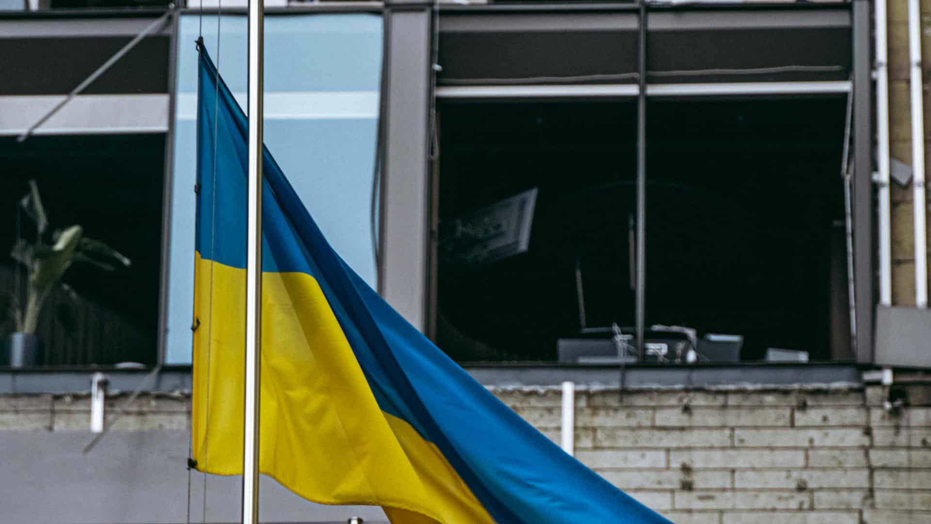 A Ukrainian flag flies from a flagpole in front of a building damaged by bombing during the Russian war in Kyiv. In the background, broken glass, walls blackened by fire and the remains of office furniture.