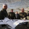 Grupo de personas, algunas con uniforme militar, entre ellas Vladimir Putin y Andrei Belousov (presidente y ministro de Defensa de la Federación Rusa respectivamente), inspeccionan varios modelos de drones sobre una mesa en una exhibición al aire libre cubierta con red de camuflaje. En primer plano, un modelo de avión blanco con una palabra en cirílico.
