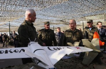Grupo de personas, algunas con uniforme militar, entre ellas Vladimir Putin y Andrei Belousov (presidente y ministro de Defensa de la Federación Rusa respectivamente), inspeccionan varios modelos de drones sobre una mesa en una exhibición al aire libre cubierta con red de camuflaje. En primer plano, un modelo de avión blanco con una palabra en cirílico.