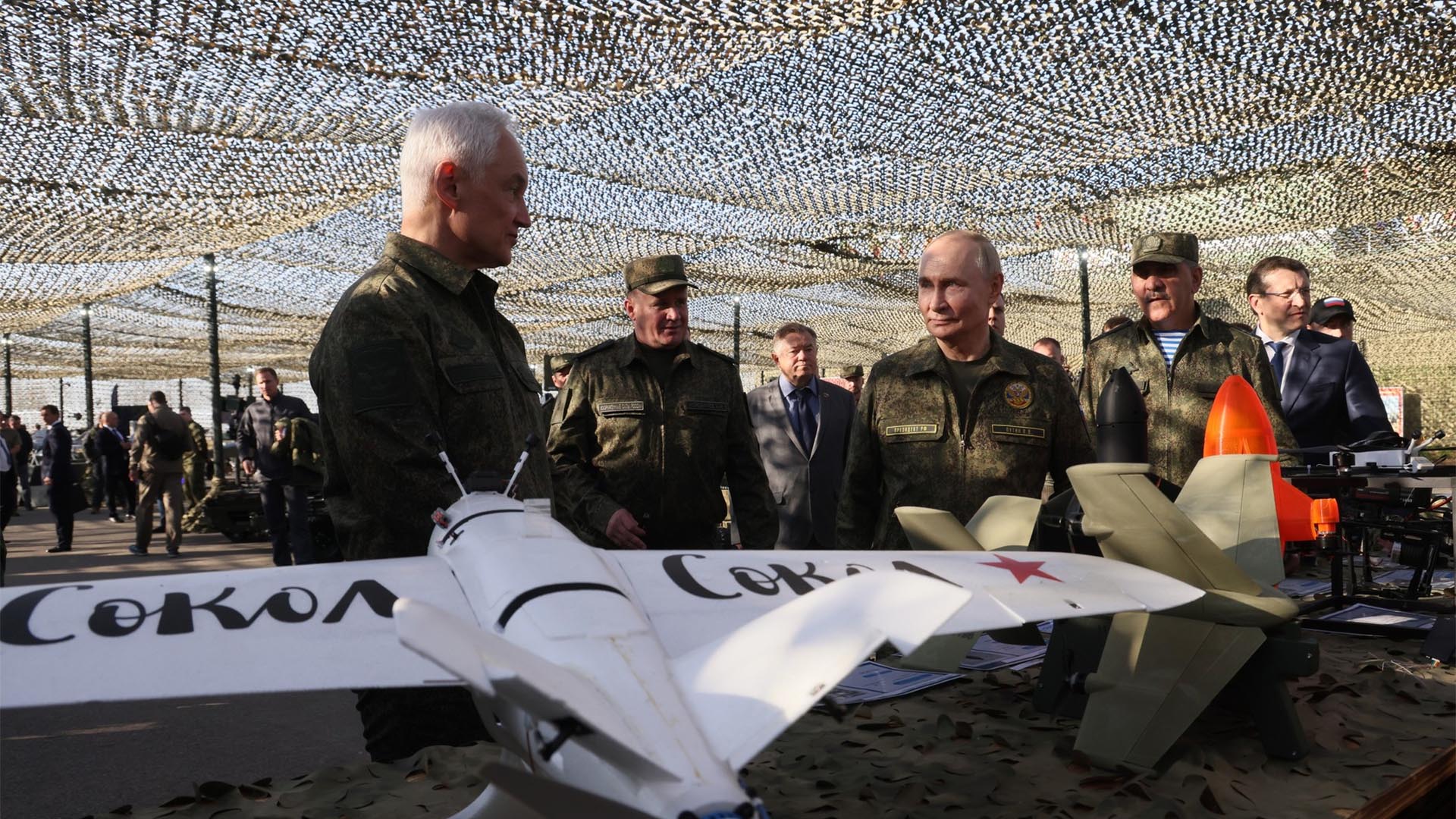 Grupo de personas, algunas con uniforme militar, entre ellas Vladimir Putin y Andrei Belousov (presidente y ministro de Defensa de la Federación Rusa respectivamente), inspeccionan varios modelos de drones sobre una mesa en una exhibición al aire libre cubierta con red de camuflaje. En primer plano, un modelo de avión blanco con una palabra en cirílico.