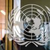 Emblema de la ONU en un cristal en la puerta deentrada del Palacio de las Naciones en Ginebra (Suiza). Detrás del cristal se ve la figura borrosa de la persona que ha tomado la foto y un pasillo con ventanales.