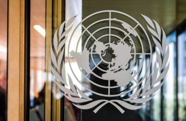 Emblema de la ONU en un cristal en la puerta deentrada del Palacio de las Naciones en Ginebra (Suiza). Detrás del cristal se ve la figura borrosa de la persona que ha tomado la foto y un pasillo con ventanales.