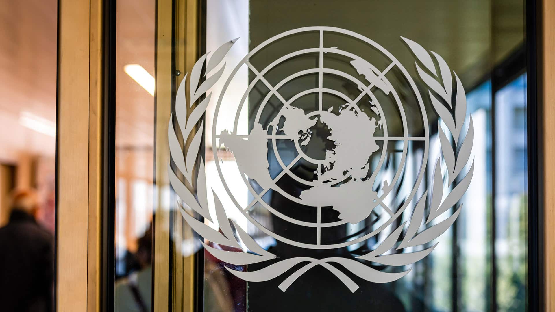 Emblema de la ONU en un cristal en la puerta deentrada del Palacio de las Naciones en Ginebra (Suiza). Detrás del cristal se ve la figura borrosa de la persona que ha tomado la foto y un pasillo con ventanales.