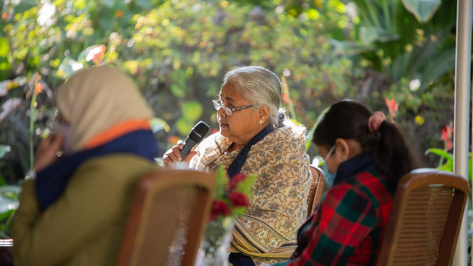 Sushila Karki, exmagistrada y primera ministra interina de Nepal elegida vía Discord, interviene en la Cumbre por la Democracia organizada por la embajada de EEUU en Katmandú en 2021. Está sentada con un micrófono en mano al lado de otras dos mujeres que están fuera de foco. Al fondo, el jardín del lugar del evento.