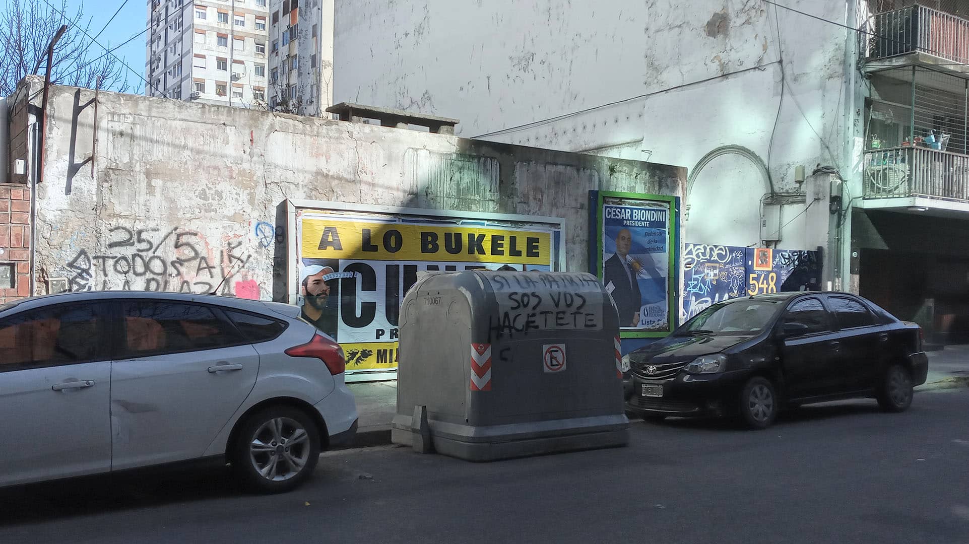 Propaganda electoral y graffitis en una calle de Buenos Aires (Argentina) en 2023. En primer plano, coches aparcados y al centro un container de basura con la leyenda “Si la patria sos vos hacete cargo”. En la pared de detrás, un cartel de Santiago Cúneo, candidato presidencial que promete una Argentina “a lo Bukele”, y a la derecha el cartel de Alejandro Biondini (h), candidato del partido neonazi Bandera Vecinal.