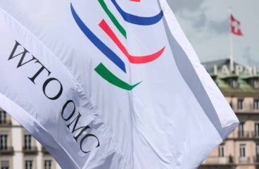 Bandera blanca con el logotipo de la Organización Mundial del Comercio (OMC) en su sede de Ginebra (Suiza). El edificio tiene la palabra «La Paix» en el tejado y una bandera suiza ondeando.