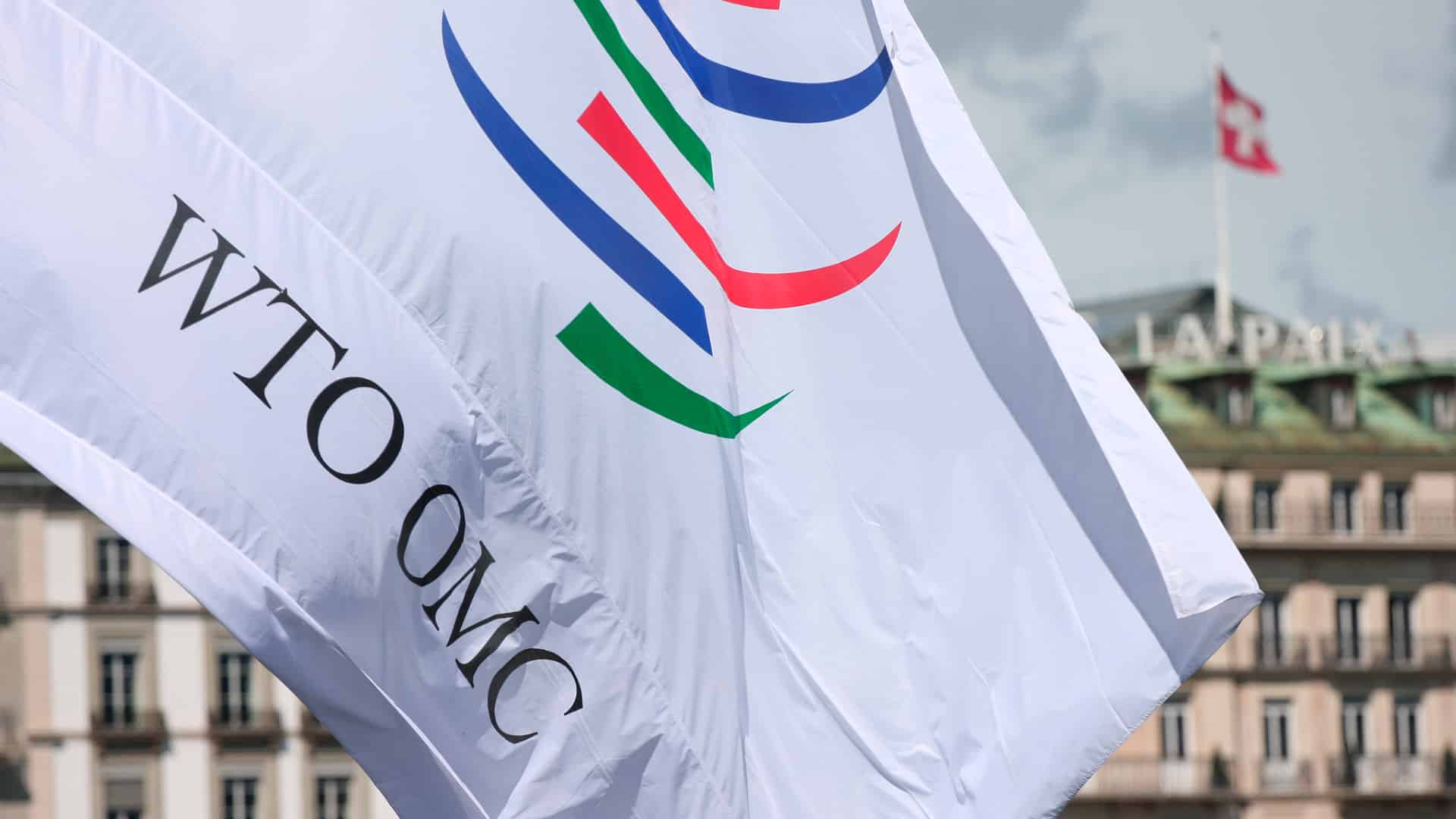 Bandera blanca con el logotipo de la Organización Mundial del Comercio (OMC) en su sede de Ginebra (Suiza). El edificio tiene la palabra «La Paix» en el tejado y una bandera suiza ondeando.