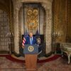Donald Trump de pie detrás de un podio con el sello presidencial de EEUU, responde a preguntas tras firmar órdenes ejecutivas en una sala de su complejo de Mar-a-Lago en Palm Beach, decorada con detalles dorados, escudo heráldico, banderas estadounidenses y mobiliario antiguo.