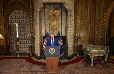 Donald Trump de pie detrás de un podio con el sello presidencial de EEUU, responde a preguntas tras firmar órdenes ejecutivas en una sala de su complejo de Mar-a-Lago en Palm Beach, decorada con detalles dorados, escudo heráldico, banderas estadounidenses y mobiliario antiguo.