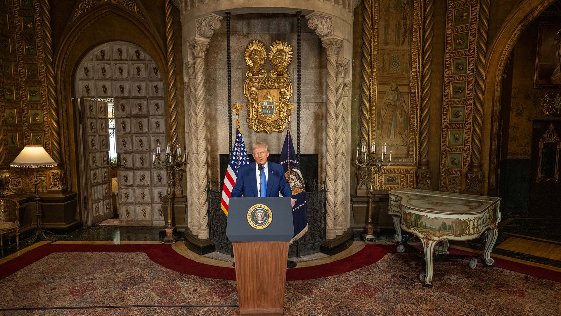 Donald Trump de pie detrás de un podio con el sello presidencial de EEUU, responde a preguntas tras firmar órdenes ejecutivas en una sala de su complejo de Mar-a-Lago en Palm Beach, decorada con detalles dorados, escudo heráldico, banderas estadounidenses y mobiliario antiguo.