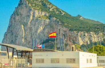 Border between Spain and Gibraltar with the Rock in the background, customs building in the foreground, and several flags flying on top (Spain, the UK, Gibraltar and the EU).