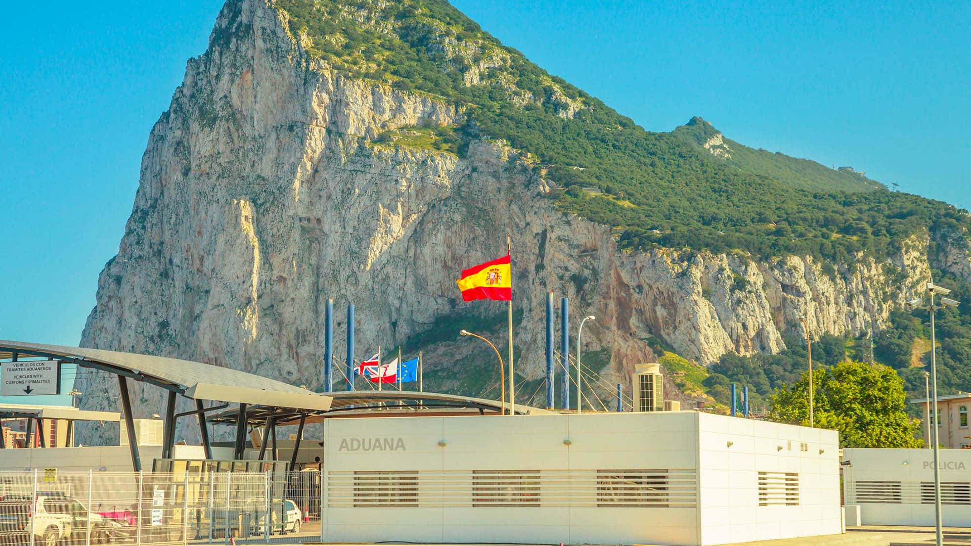 Border between Spain and Gibraltar with the Rock in the background, customs building in the foreground, and several flags flying on top (Spain, the UK, Gibraltar and the EU).