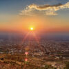 Panorámica de la ciudad de Sueida (Siria) tomada en 2012 desde la colina de Ahmar al atardecer, con el sol en el horizonte, campos verdes y cielo con tonos anaranjados y azul oscuro.