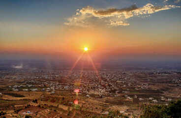 Panorámica de la ciudad de Sueida (Siria) tomada en 2012 desde la colina de Ahmar al atardecer, con el sol en el horizonte, campos verdes y cielo con tonos anaranjados y azul oscuro.
