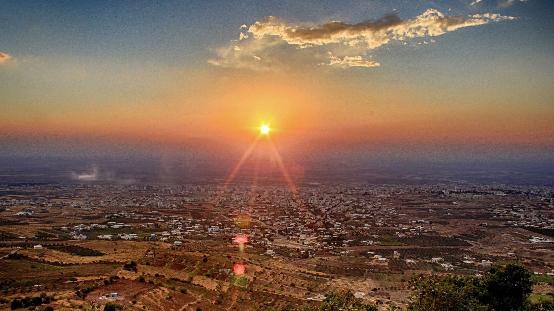 Panorámica de la ciudad de Sueida (Siria) tomada en 2012 desde la colina de Ahmar al atardecer, con el sol en el horizonte, campos verdes y cielo con tonos anaranjados y azul oscuro.