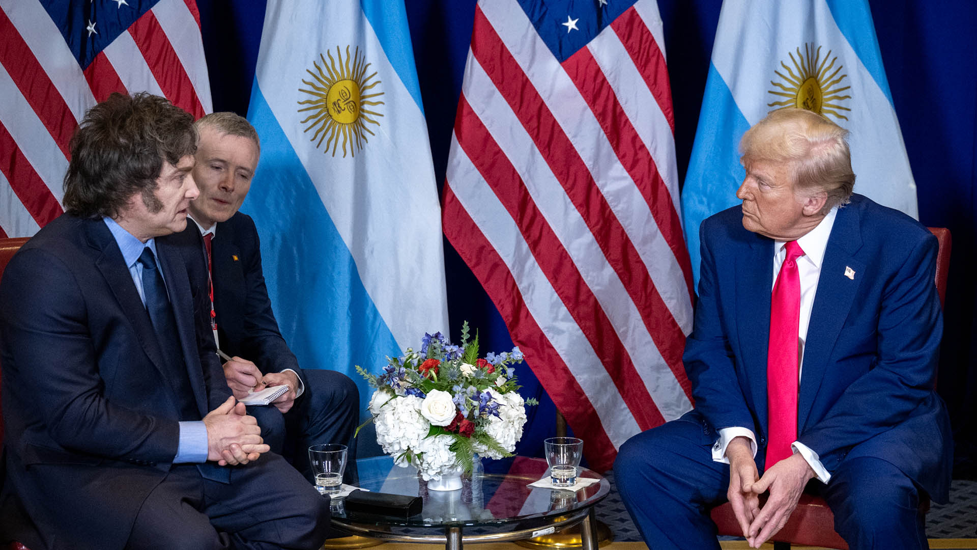 Javier Milei, su traductor, y Donald Trump durante el encuentro entre ambos mandatarios en el Gaylord National Resort & Convention Center de Oxon Hill, Maryland (22/02/2025). Al fondo, las banderas de Argentina y Estados Unido y un arreglo floral sobre la mesa en el centro.