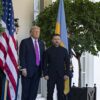 Donald Trump (a la izquierda) recibe a Volodímir Zelenski (a la derecha) en la entrada del ala oeste de la Casa Blanca el 17 de octubre de 2025. Les flanquean dos guardias de honor y las banderas de EEUU y Ucrania, además de arbustos en macetas. En primer plano a la derecha, la parte delantera de un vehículo con un banderín estadounidense.
