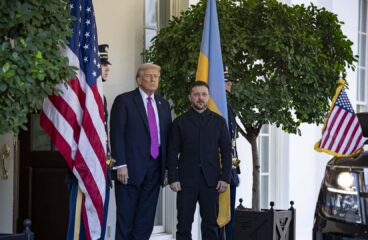 Donald Trump (a la izquierda) recibe a Volodímir Zelenski (a la derecha) en la entrada del ala oeste de la Casa Blanca el 17 de octubre de 2025. Les flanquean dos guardias de honor y las banderas de EEUU y Ucrania, además de arbustos en macetas. En primer plano a la derecha, la parte delantera de un vehículo con un banderín estadounidense.
