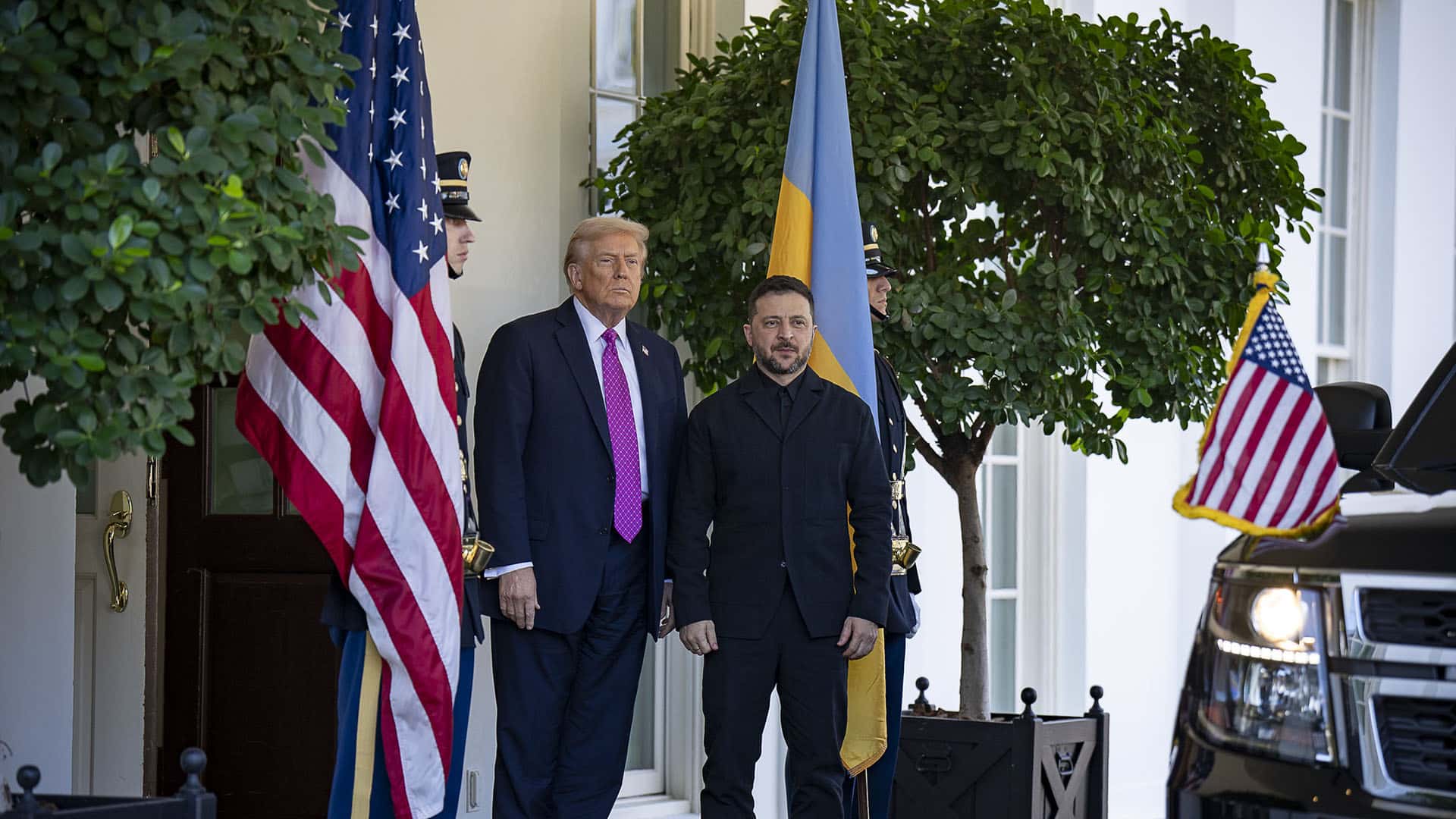 Donald Trump (a la izquierda) recibe a Volodímir Zelenski (a la derecha) en la entrada del ala oeste de la Casa Blanca el 17 de octubre de 2025. Les flanquean dos guardias de honor y las banderas de EEUU y Ucrania, además de arbustos en macetas. En primer plano a la derecha, la parte delantera de un vehículo con un banderín estadounidense.