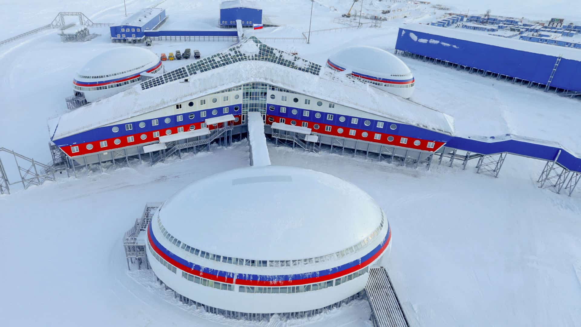 Vista aérea de la base militar Arctic Trefoil de la Federación Rusa en la isla Alexandra Land en el Ártico, con edificios circulares y rectangulares pintados en azul, rojo y blanco, rodeados de nieve.