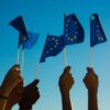 Brazos levantados sosteniendo banderitas de la Unión Europea hacia el cielo azul.