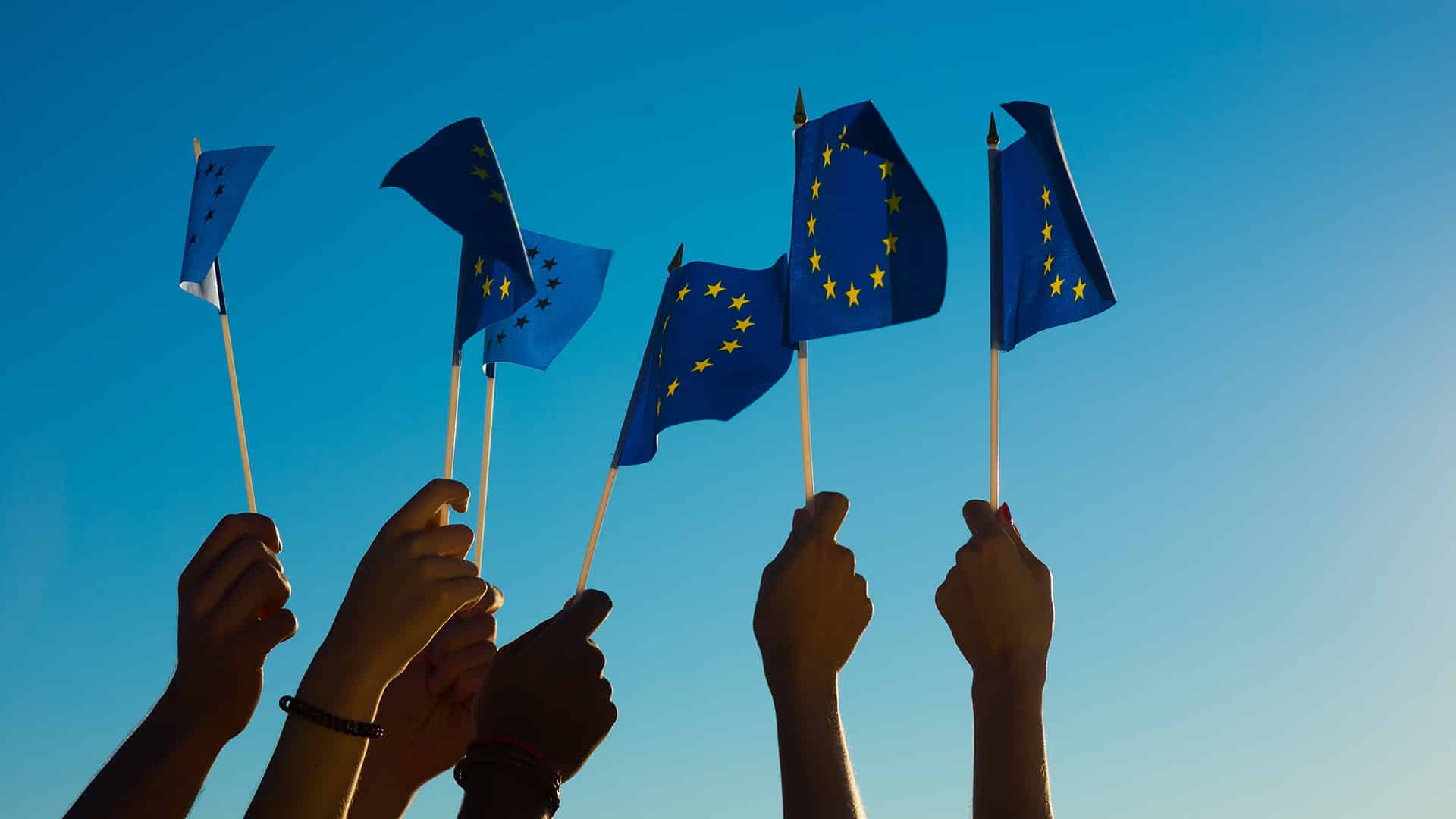 Brazos levantados sosteniendo banderitas de la Unión Europea hacia el cielo azul.