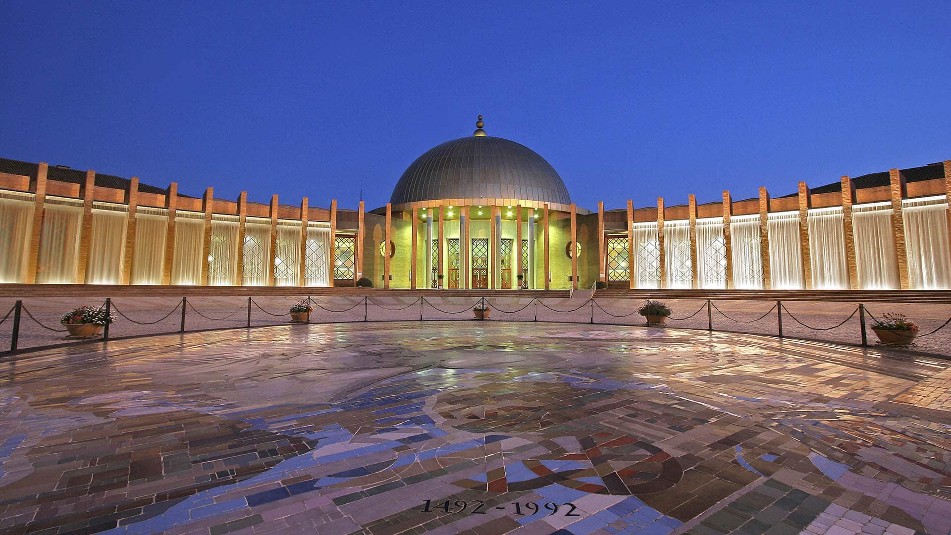 Plaza exterior y vista frontal del Palacio de Exposiciones y Congresos de Sevilla, con estructura semicircular, la cúpula central iluminada y suelo decorado con mosaicos que incluyen la inscripción “1492-1992”.