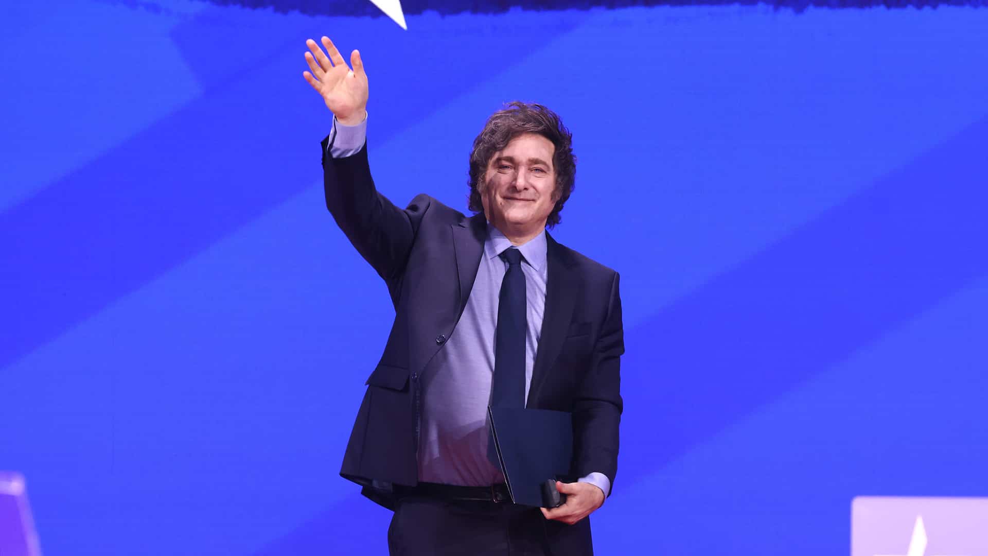 Javier Milei, con traje oscuro, camisa violeta y una carpeta en su mano izquierda, saluda al público desde el escenario de la Conferencia de Acción Política Conservadora (CPAC) en marzo de 2025.