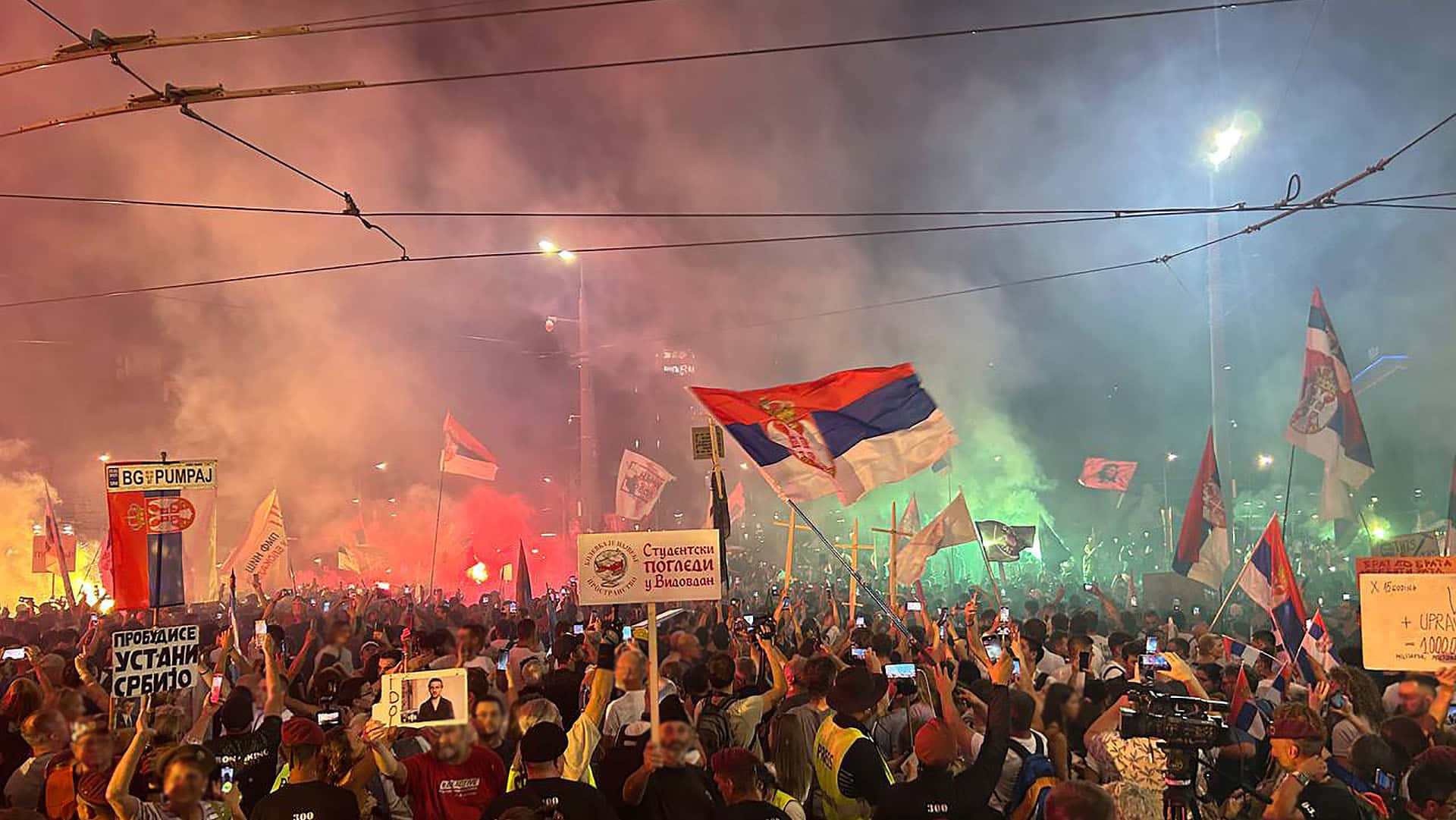 Gran protesta nocturna llamada «Nos vemos en Vidovdan», con multitud de personas portando banderas de Serbia, pancartas y teléfonos móviles, rodeados de humo y luces de colores.