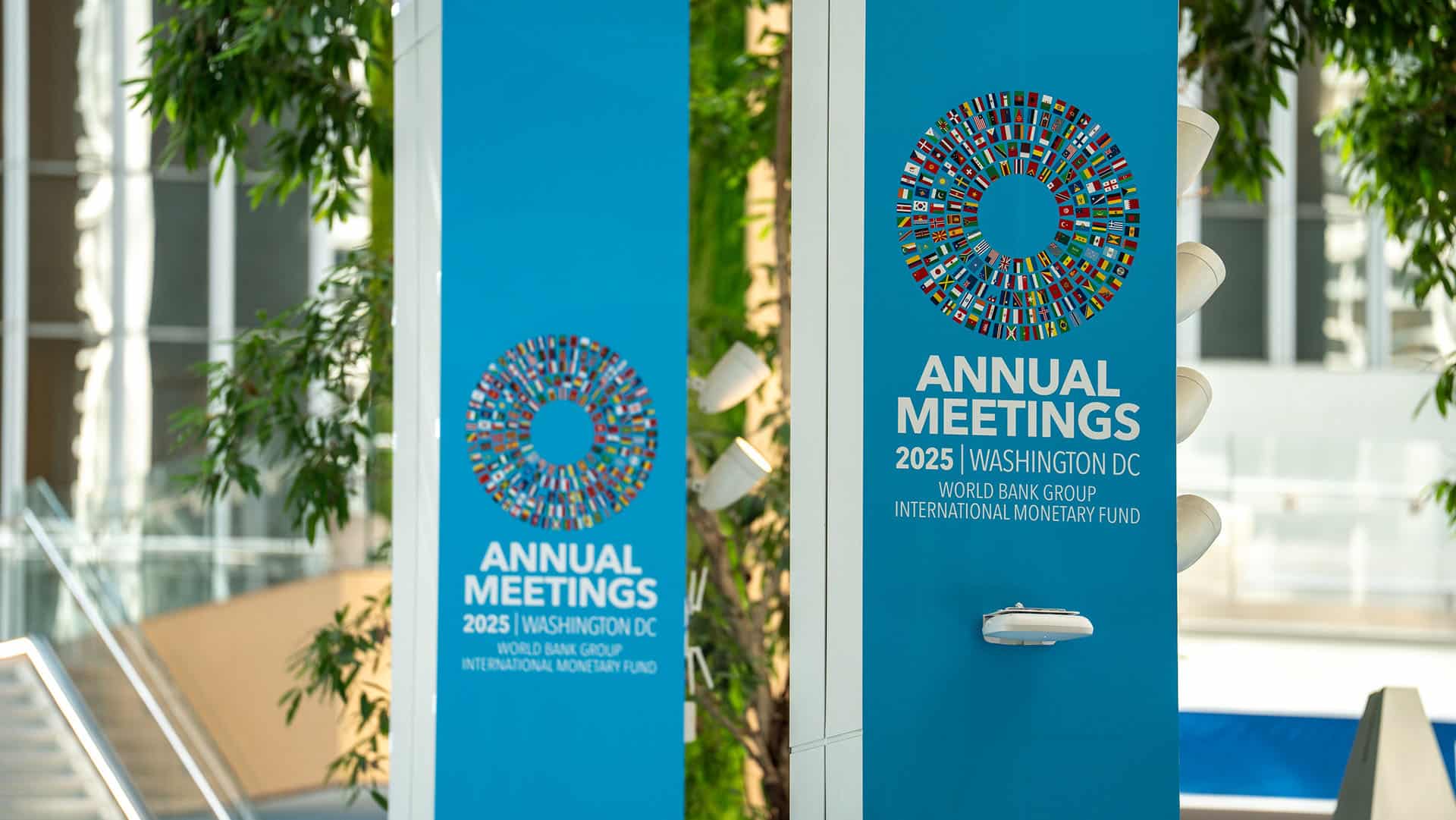 Paneles con el logotipo y texto de las Reuniones Anuales 2025 en Washington D.C. del Banco Mundial y el Fondo Monetario Internacional, decorados con un círculo formado por banderas de distintos países.