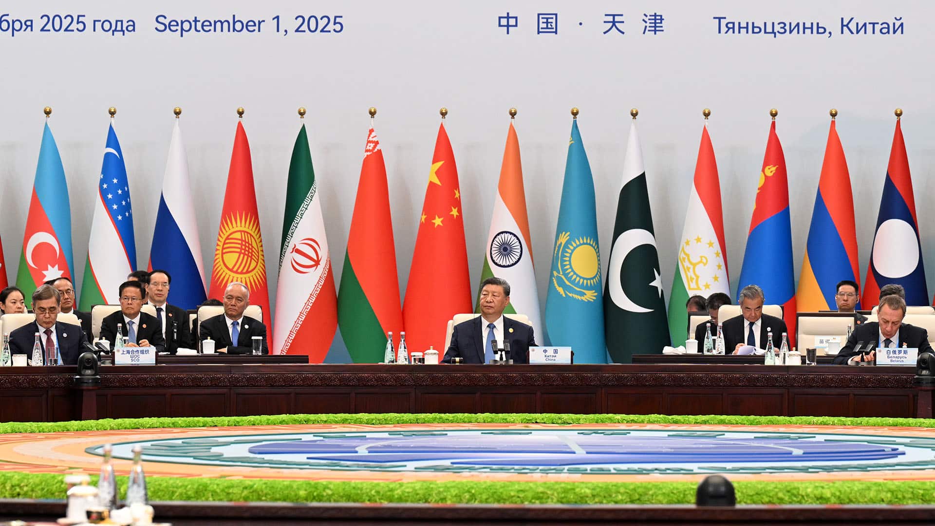 Mesa principal del plenario de la 25ª cumbre de jefes de Estado ampliada de la Organización de Cooperación de Shanghái (OCS) en Tianjin, China. Al centro, Xi Jinping (presidente de China) flanqueado por el resto de representantes y delegaciones. Al fondo, las banderas de los países miembros de la OCS, con un texto que indica la fecha (1 de septiembre de 2025) y la ubicación (Tianjin, China).