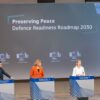 Rueda de prensa sobre la Hoja de ruta para la Preparación en materia de defensa 2030 de la Comisión Europea. Dos hombres y dos mujeres hablan desde sus respectivos atriles ante un auditorio con personas. Al fondo, una pantalla que proyecta imágenes de equipo military y un satélite con las palabras “Preserving Peace. Defence Readiness Roadmap 2023”. De izquierda a derecha: Thomas Regnier (portavoz sobre soberanía digital, defensa, espacio e investigación), Henna Virkkunen y Kaja Kallas (vicepresidentas de la Comisión), y Andrius Kubilius (comisari europeo).