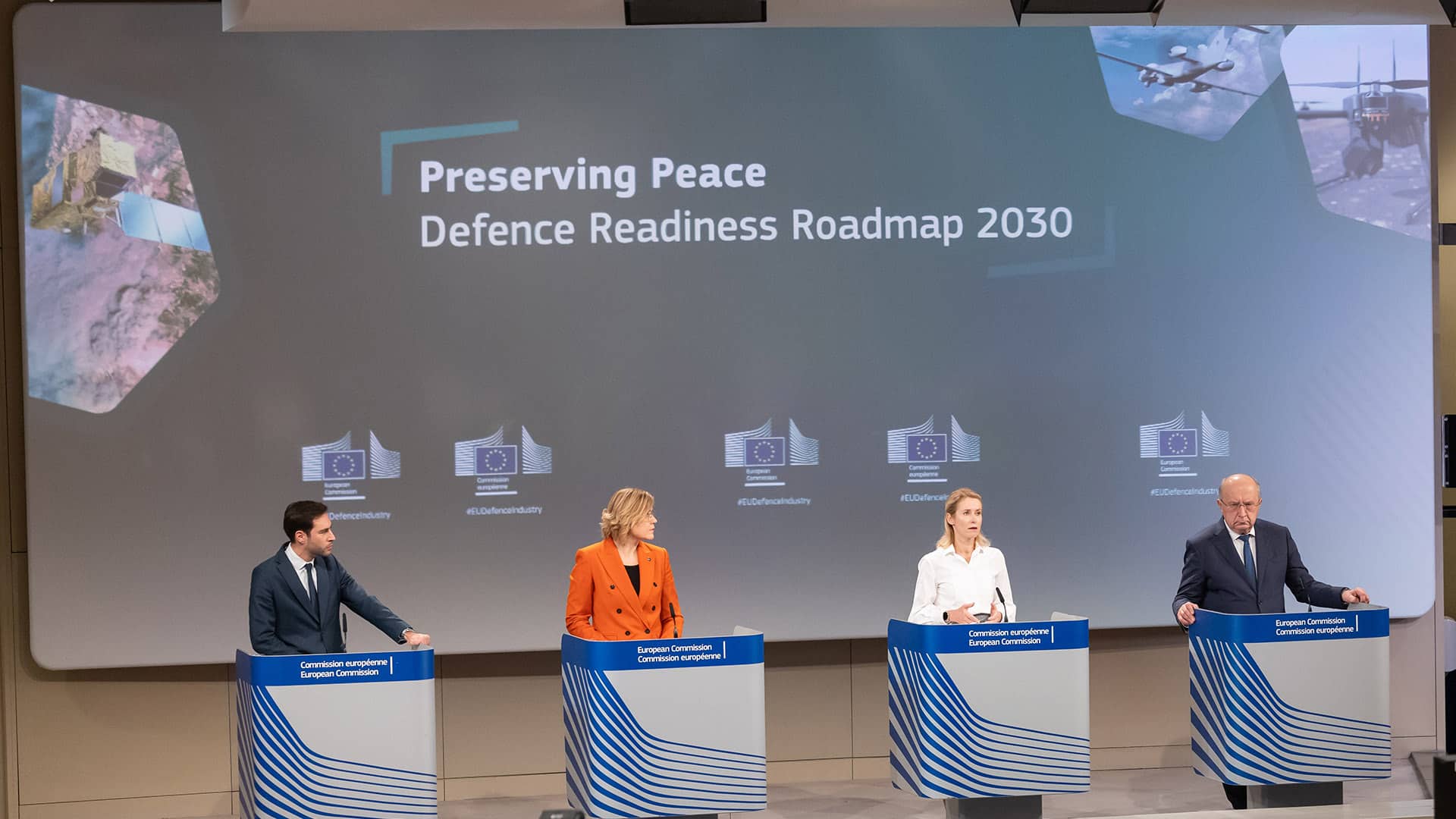 Rueda de prensa sobre la Hoja de ruta para la Preparación en materia de defensa 2030 de la Comisión Europea. Dos hombres y dos mujeres hablan desde sus respectivos atriles ante un auditorio con personas. Al fondo, una pantalla que proyecta imágenes de equipo military y un satélite con las palabras “Preserving Peace. Defence Readiness Roadmap 2023”. De izquierda a derecha: Thomas Regnier (portavoz sobre soberanía digital, defensa, espacio e investigación), Henna Virkkunen y Kaja Kallas (vicepresidentas de la Comisión), y Andrius Kubilius (comisari europeo).
