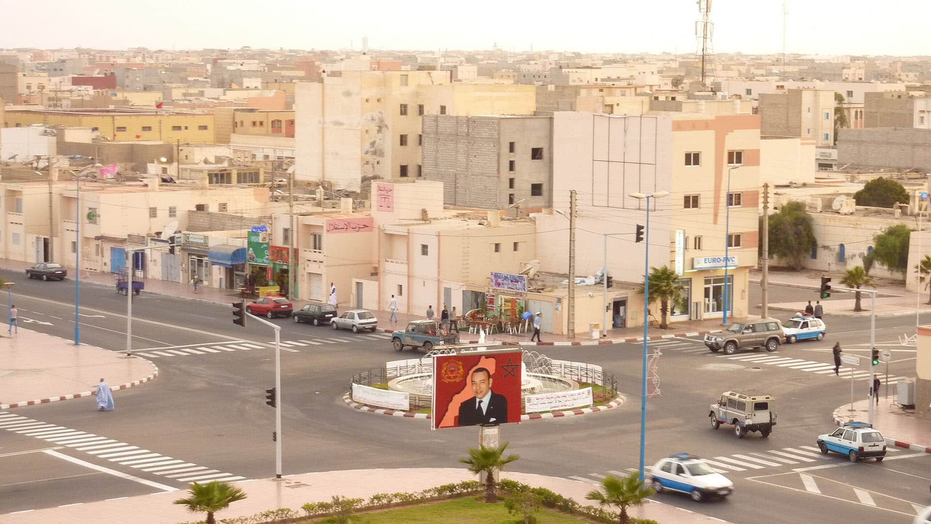 Vista aérea de una intersección en Dajla (ciudad del Sáhara Occidental) con edificios bajos, tráfico moderado y una rotonda central decorada con un gran cartel que muestra un retrato oficial del Rey Mohamed VI de Marruecos, en un entorno urbano desértico.