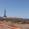 Plataforma de perforación petrolera no convencional en Vaca Muerta (Argentina), en un terreno árido con vegetación baja, una tubería naranja en primer plano y el lago Mari Menuco visible en el horizonte bajo un cielo despejado.