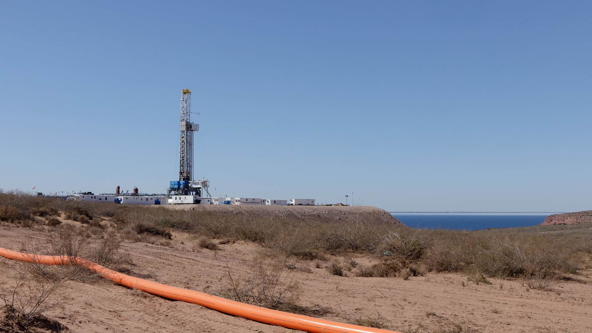 Plataforma de perforación petrolera no convencional en Vaca Muerta (Argentina), en un terreno árido con vegetación baja, una tubería naranja en primer plano y el lago Mari Menuco visible en el horizonte bajo un cielo despejado.