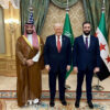 Mohamed bin Salman, Donald Trump y Ahmed al-Sharaa posando en una sala decorada con cortinas verdes, alfombra ornamental y lámparas doradas, con banderas de Estados Unidos, Arabia Saudí y Siria al fondo. La foto se hizo durante la visita de Estado de Trump a Arabia Saudí en mayo de 2025.