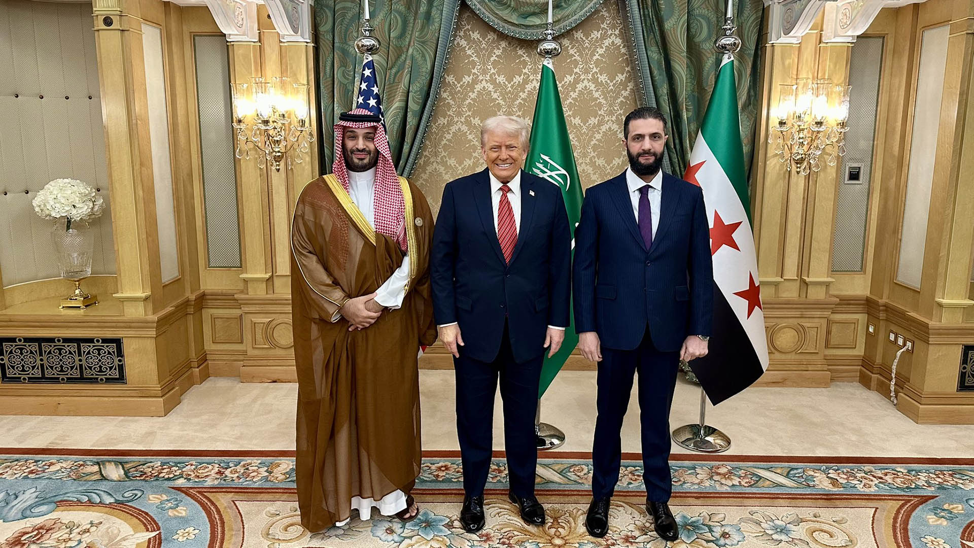 Mohamed bin Salman, Donald Trump y Ahmed al-Sharaa posando en una sala decorada con cortinas verdes, alfombra ornamental y lámparas doradas, con banderas de Estados Unidos, Arabia Saudí y Siria al fondo. La foto se hizo durante la visita de Estado de Trump a Arabia Saudí en mayo de 2025.