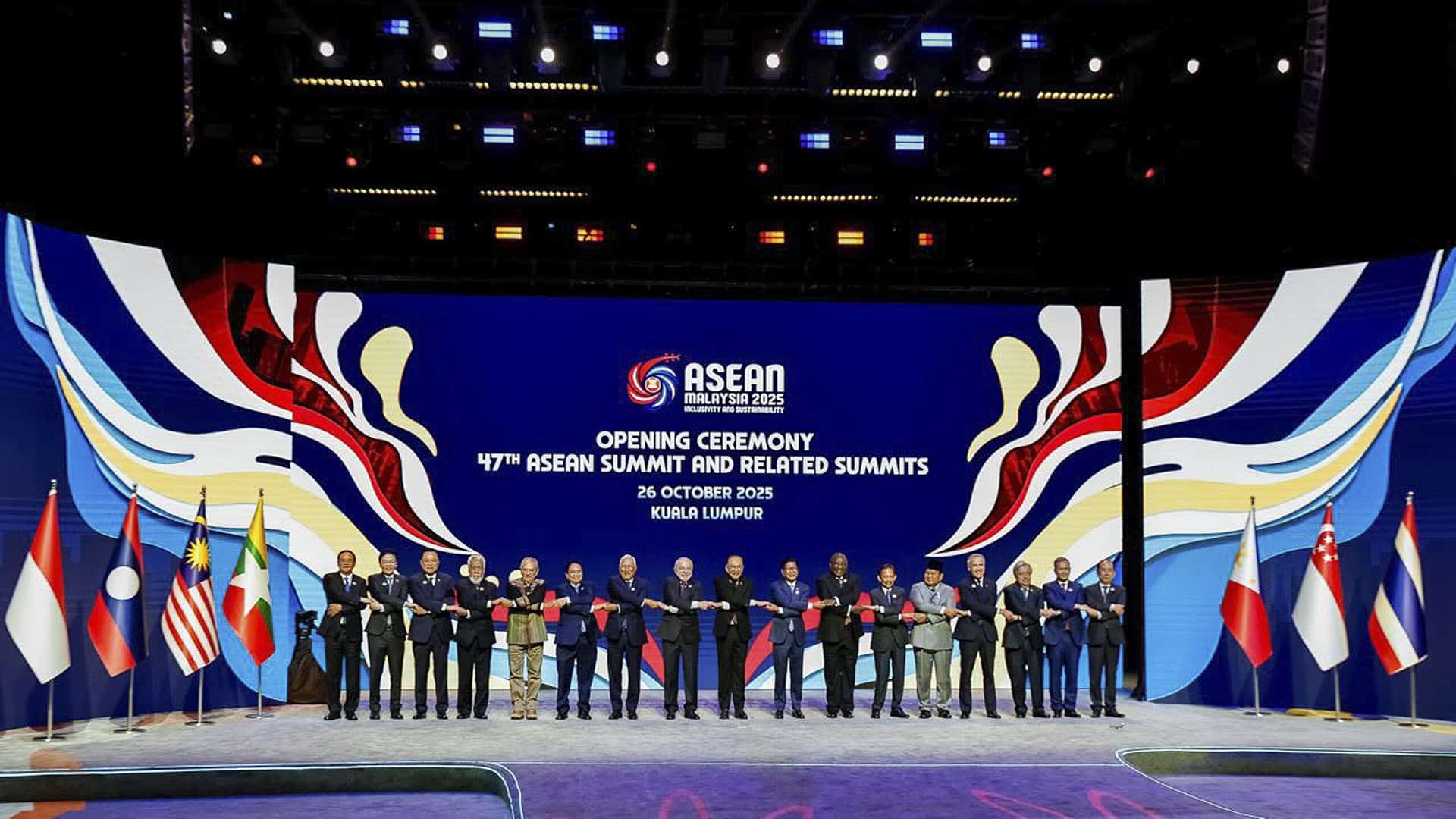 Foto de familia de la inauguración de la 47ª Cumbre de la ASEAN y cumbres relacionadas en Kuala Lumpur, Malasia. En el escenario, los jefes de Estado y Gobierno de los países con las manos entrelazadas, flanqueados por banderas y al fondo pantallas con el logo de la cumbre y figuras abstractas.
