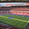 Vista panorámica del Estadio Metropolitano durante un partido, con gradas llenas de aficionados y el césped preparado para el encuentro, mostrando el nombre Atlético de Madrid en los asientos.
