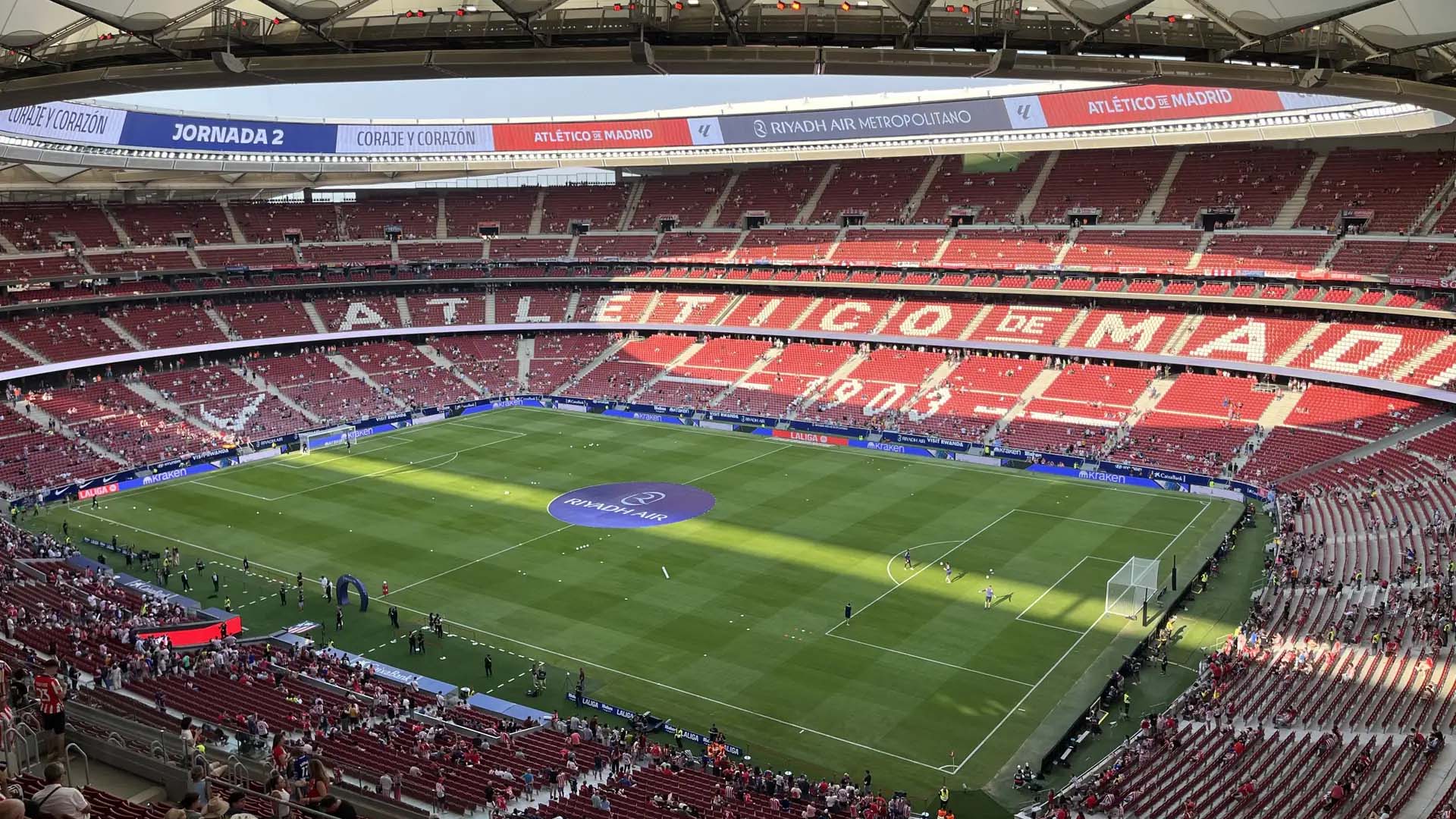 Vista panorámica del Estadio Metropolitano durante un partido, con gradas llenas de aficionados y el césped preparado para el encuentro, mostrando el nombre Atlético de Madrid en los asientos.