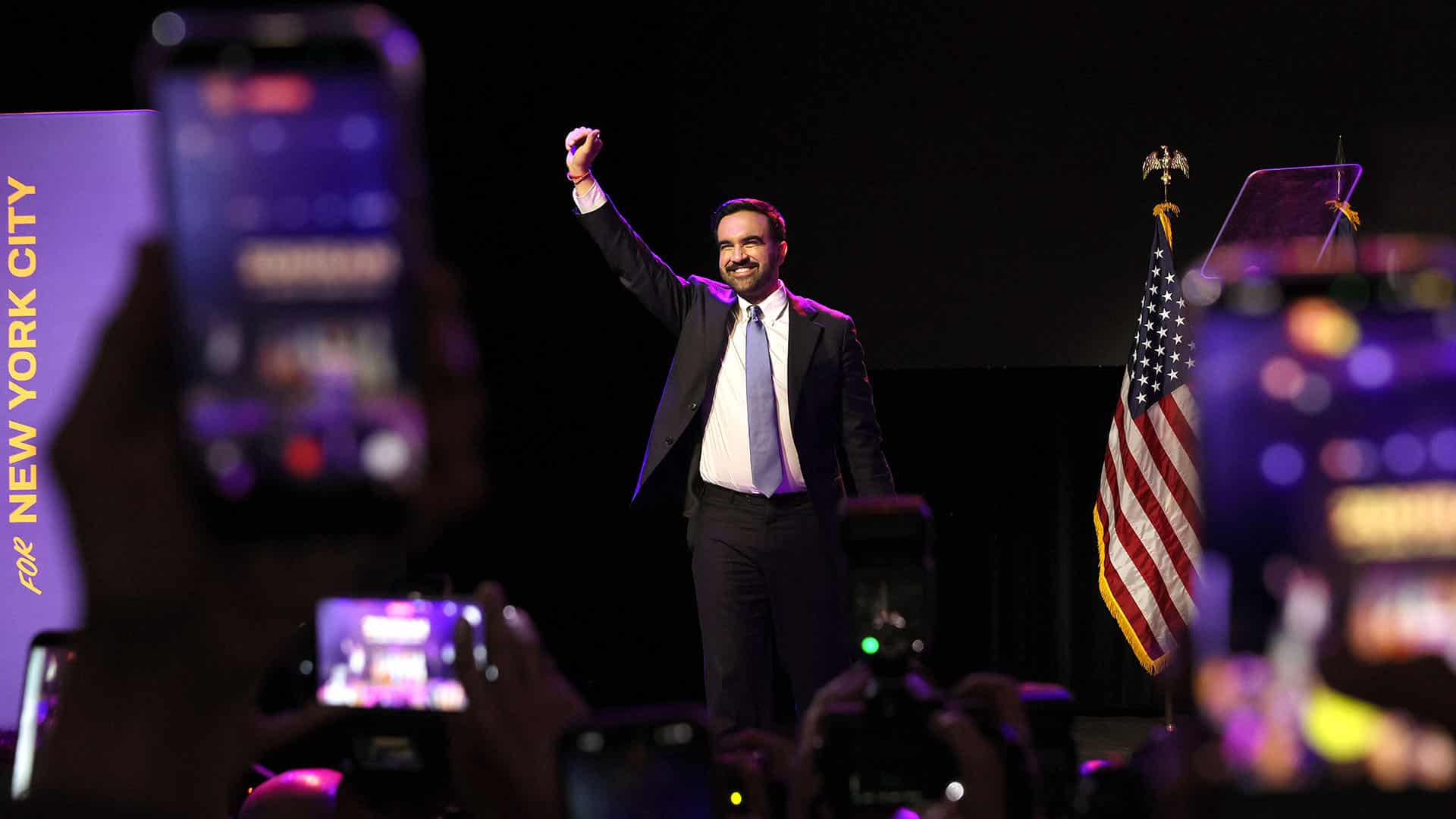 Zohran Mamdani celebra su victoria en el escenario con el brazo en alto durante la fiesta electoral celebrada en el Brooklyn Paramount, en el distrito de Brooklyn de la ciudad de Nueva York (4/11/202). A un lado, la bandera de Estados Unidos en un mástil, y al otro parte de un cartel lila con las palabras “For New York City”.
