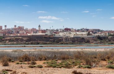 Vista panorámica de la ciudad de El Aaiún, en el territorio del Sáhara Occidental. En primer plano, un terreno árido con vegetación dispersa; más atrás, una superficie extensa de agua; y, al fondo, una vista general de la ciudad de El Aaiún.